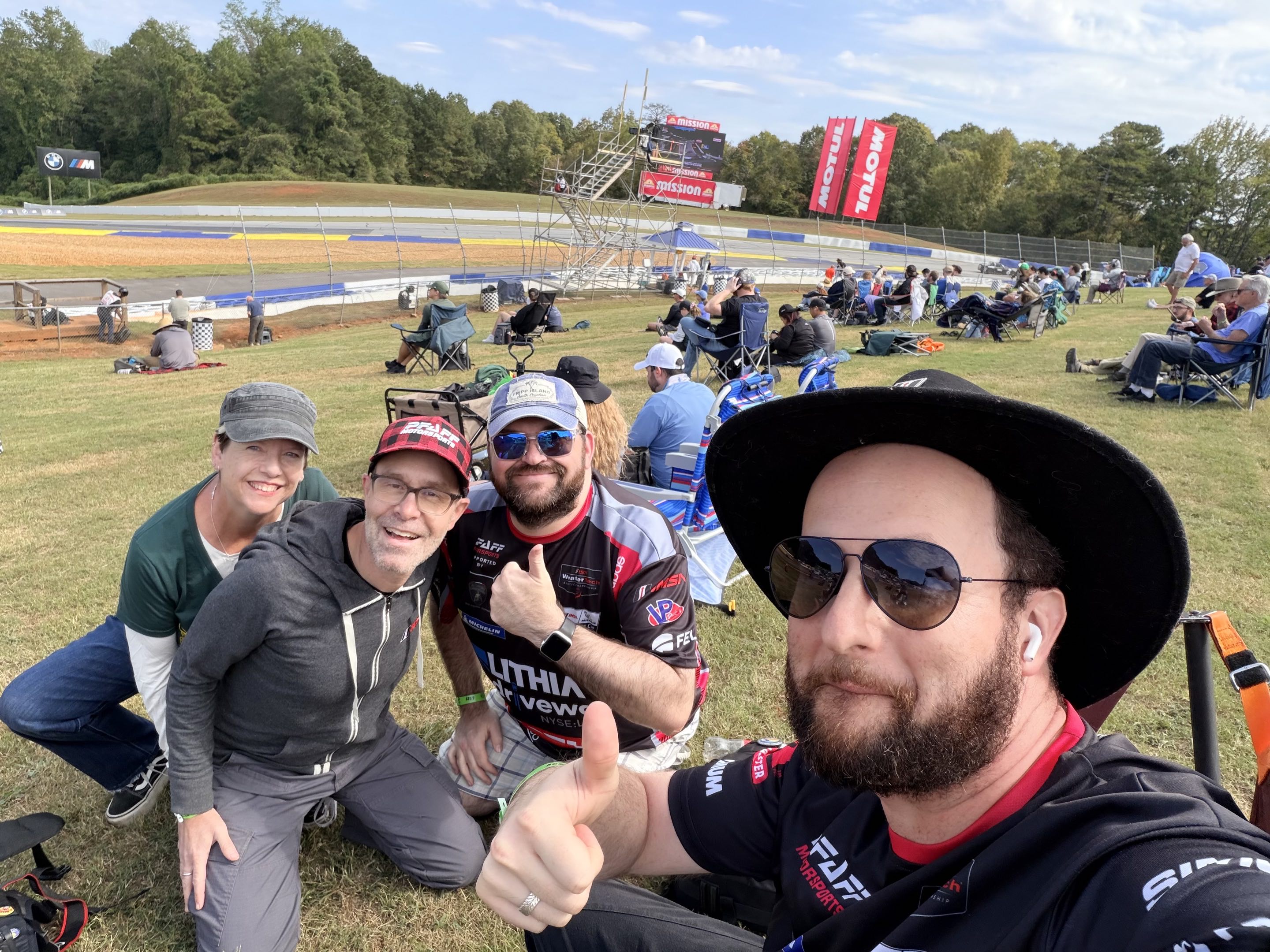 A selfie of people smiling and giving thumbs up by a race track on a beautiful day